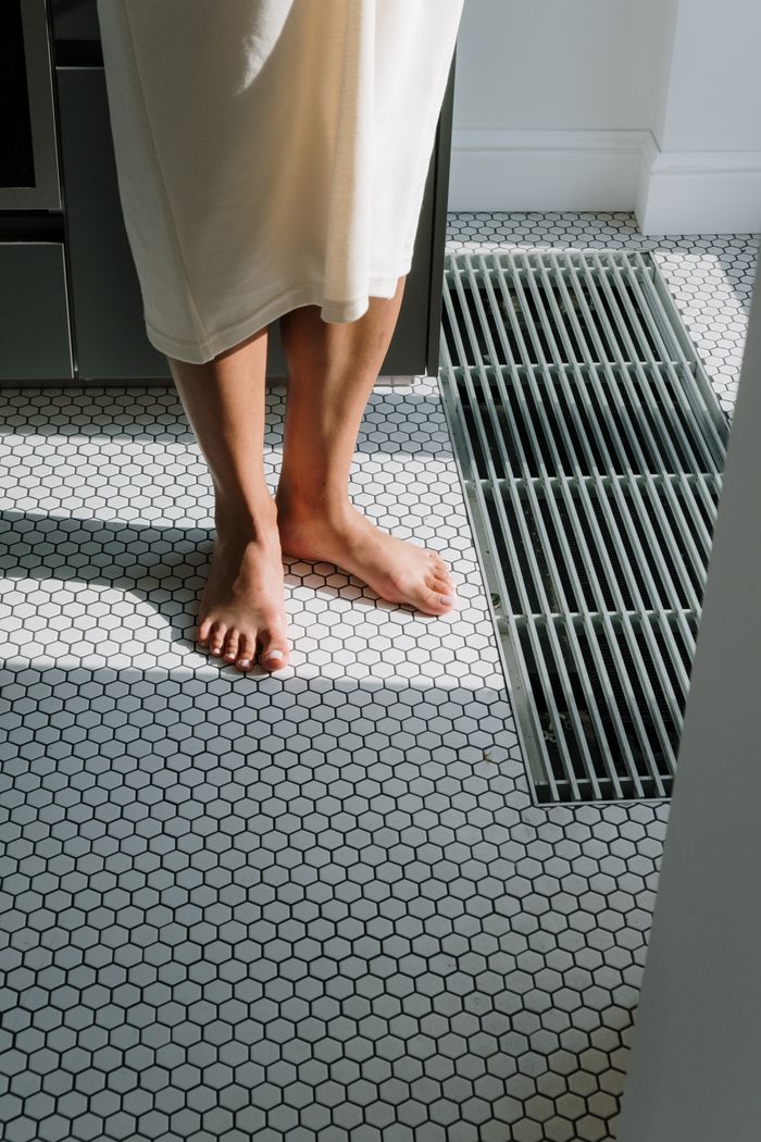 services-01 A woman stands barefoot on a sunlit hexagonal tiled floor indoors. Elegant and serene.